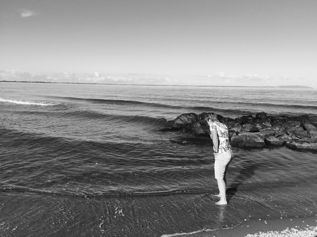 Et sort/hvidt foto af en kvinde, der står i vandkanten og kigger ned i vandet.