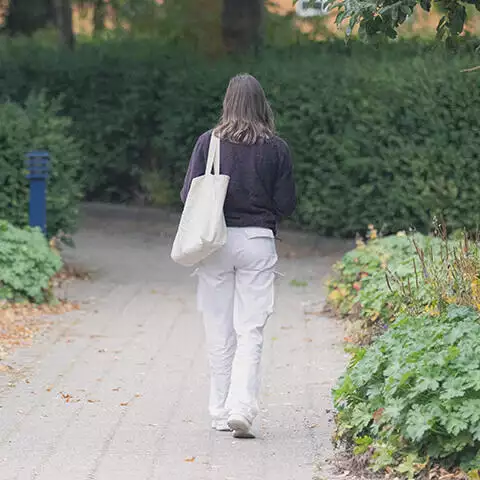 En ung kvinde går ned ad en stenbelagt sti med et net over skulderen. Hun er omgivet af træer og buske